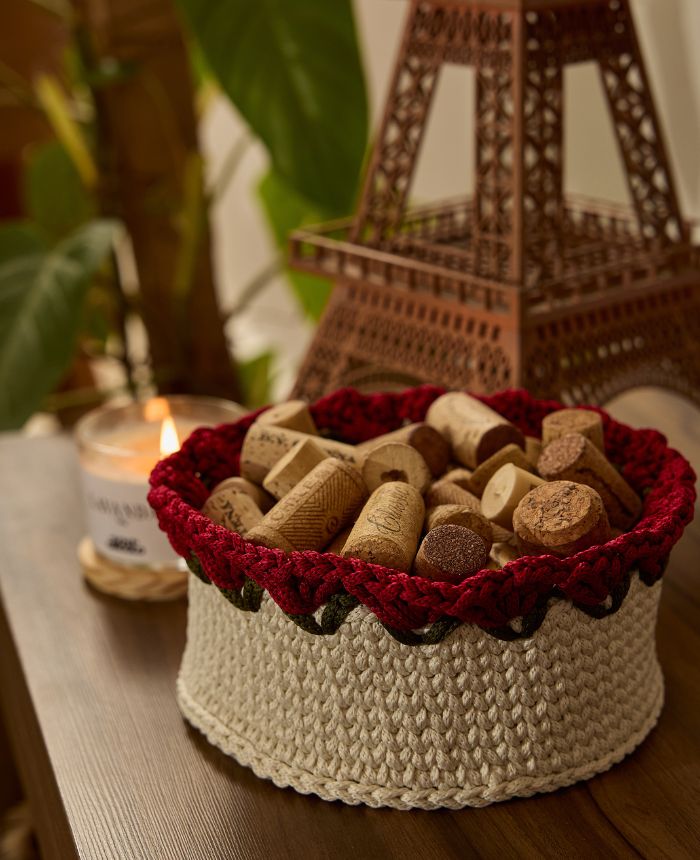 Circulo Fio Náutico Poly 5 mm – Polyester Cord Yarn ( 178 m / 500 g) - A round crocheted basket with a cream-colored body and a deep red scalloped rim, filled with cork stoppers. The basket sits on a wooden table in a cozy setting with a lit candle and a decorative Eiffel Tower in the background.