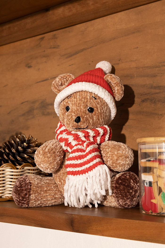 Círculo Amigurumi Pelúcia – Polyester Chenille Yarn (131 m / 85 g) - A plush teddy bear wearing a red and white knit Santa hat and matching striped scarf, sitting on a wooden shelf with a pinecone and a small jar nearby.