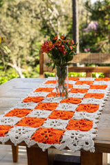Círculo Natural Cotton Multicolor 4/6 – Cotton Yarn (226 m / 200 g) – A crocheted table runner with alternating orange and white granny squares laid out on a wooden table, with a glass vase of orange flowers placed on top in an outdoor garden setting.