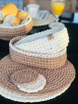 Círculo Barroco Maxcolor Sparkle – Cotton Yarn (216m / 200g) - A crocheted wide-brim sun hat in layered beige, taupe, and off-white, with a small gold-tone logo tab on the side. The hat is displayed on a dark table surface with a blurred background showing a basket of bread and a glass of orange juice.