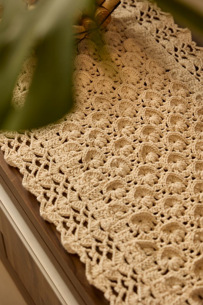 Circulo Barroco Natural Brilho  - Natural Cotton Yarn Gold/Silver, 657m / 433m, 400g - A close-up of a beige crocheted lace table runner draped over a wooden surface, with soft green leaves partially framing the top left corner.