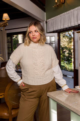 Circulo InLove  - 100% Brazilian Cotton Yarn  (125m/100g)- A woman wearing a cream cable-knit mock neck sweater vest over a white long-sleeve shirt and brown pants, standing in a home interior near a kitchen island.