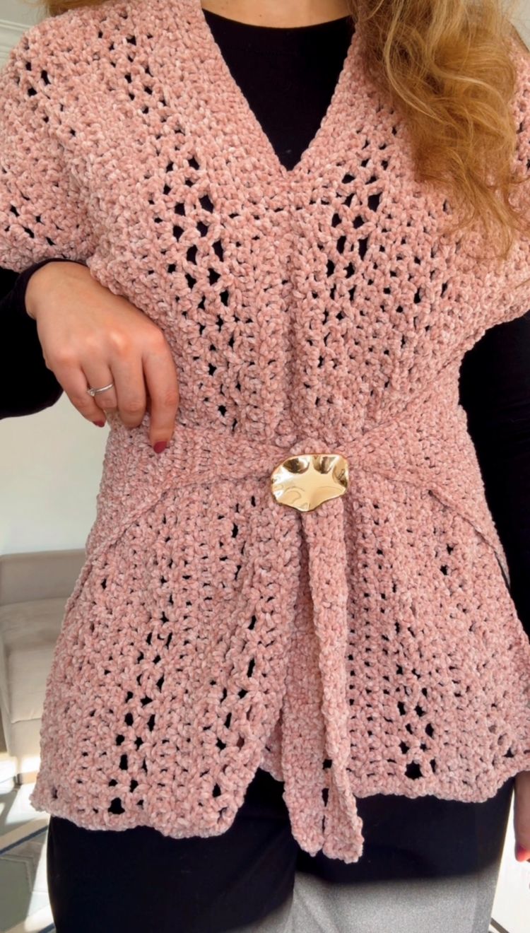Círculo Velvet – 100% Polyester Chenille Yarn (165 m / 100 g) - A close-up of a person wearing a pink crocheted wrap vest with a tie belt and a gold heart-shaped buckle, layered over a black top.