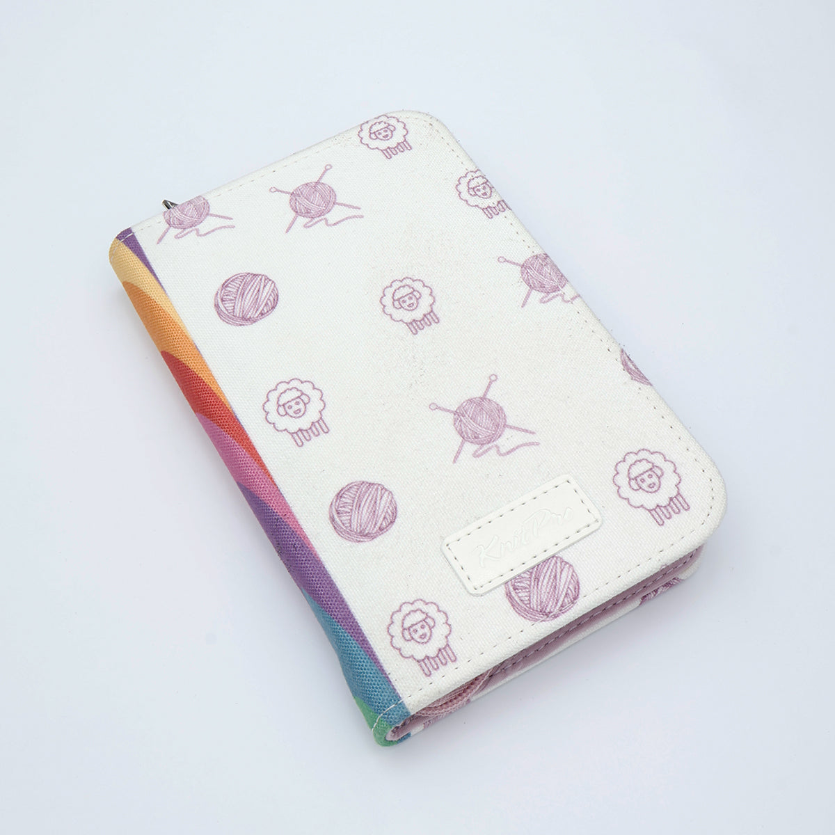 KnitPro Symfonie 2 Mini interchangeable needle set, 5 cm - A zippered notebook or planner cover with a cream-colored faux leather exterior decorated with a pattern of purple yarn balls and sheep, and a colorful striped edge showing rainbow hues.