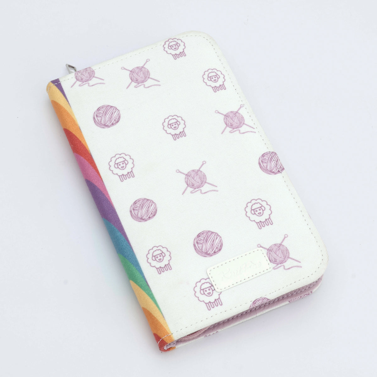 KnitPro Symfonie 2 Special interchangeable needle set, 10 cm, Deluxe - A zippered wallet or notebook cover in off-white faux leather, decorated with a repeating pattern of purple yarn balls and sheep, with a vibrant rainbow-colored fabric spine.